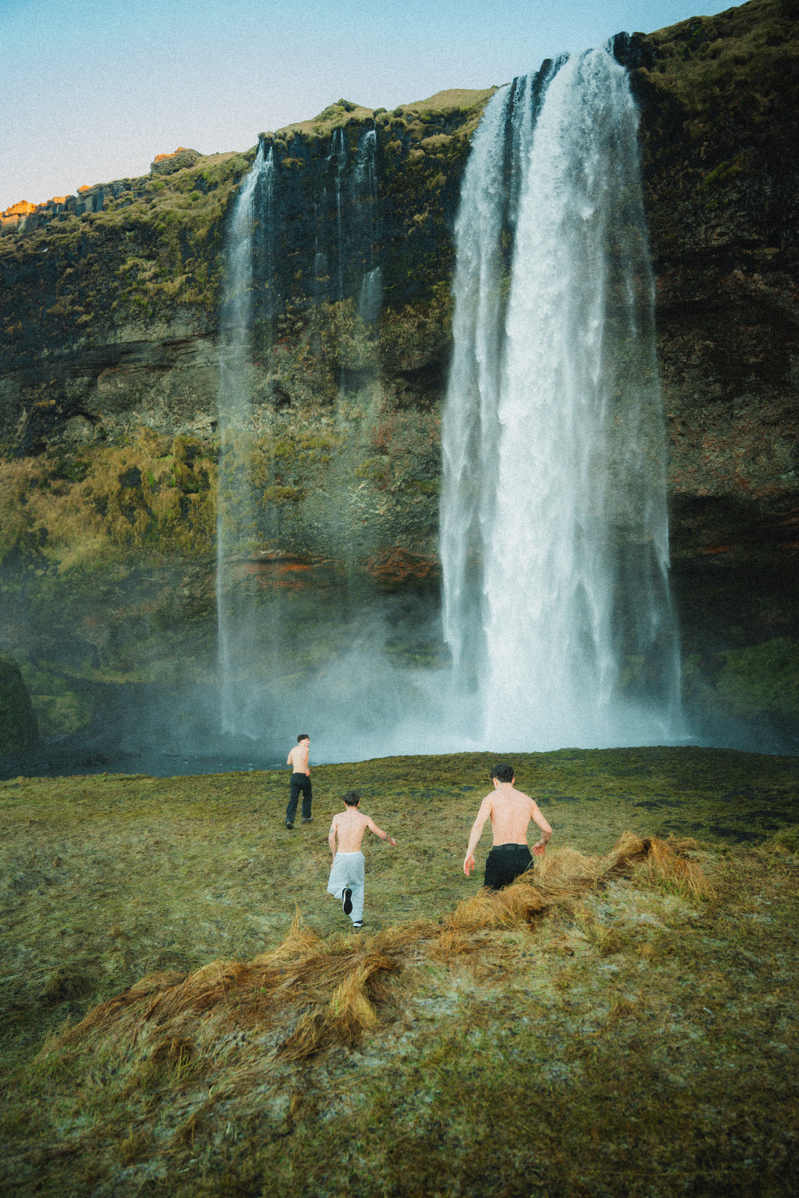 Três amigos correm em direção a uma grande cascata na Islândia